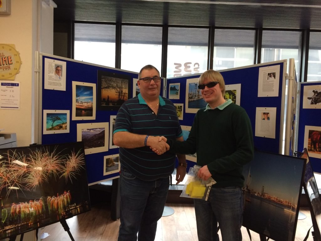 David Shervill handing the Memory Sticks to Dave Taylor in Portsmouth Central Library, Guildhall Square, Portsmouth. They are standing in front of some of the photographs on display.