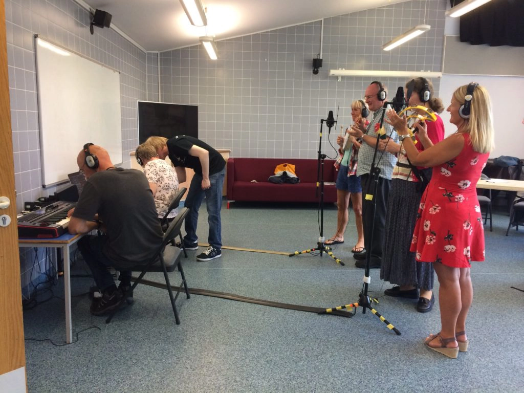 Included is a picture of the participants. One is sitting in front of the laptop recording the group, whilst another is playing the MIDI Keyboard. The other group members are standing behind microphones, playing percussion instruments and singing.