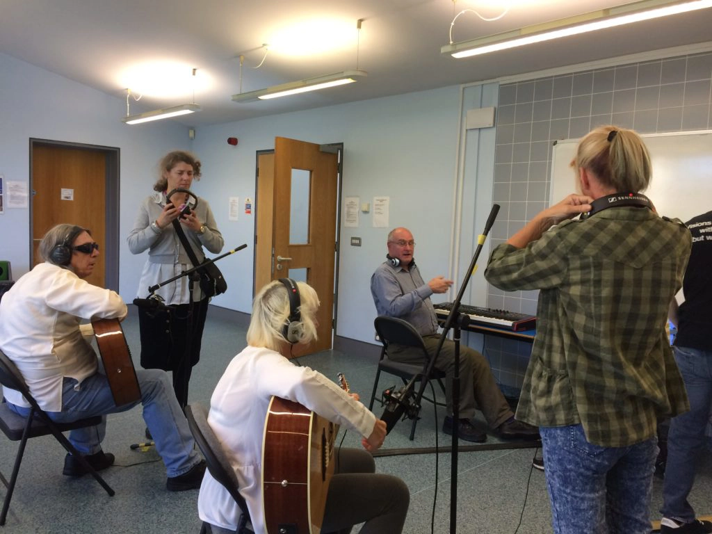 Included is a picture of the group of five discussing ideas for recording their piece of music. One participant is sitting behind a MIDI keyboard, whilst two others are sitting behind a microphone each playing acoustic guitars. Two other people are standing by each microphone, ready to change the position of the microphone if necessary.