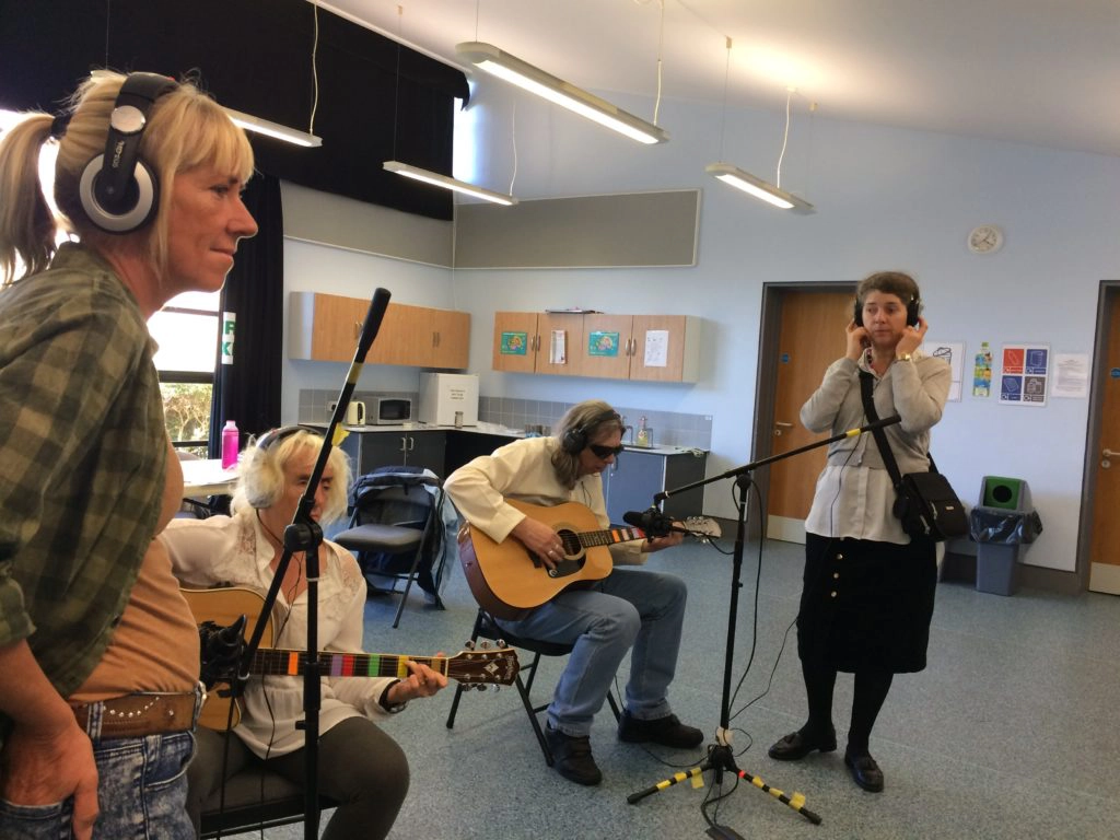 Included is a picture of four participants. Two are sitting behind a microphone playing acoustic guitars, and two others are standing by each microphone, ready to change the position if needed. All the participants are wearing headphones to listen to the recording.