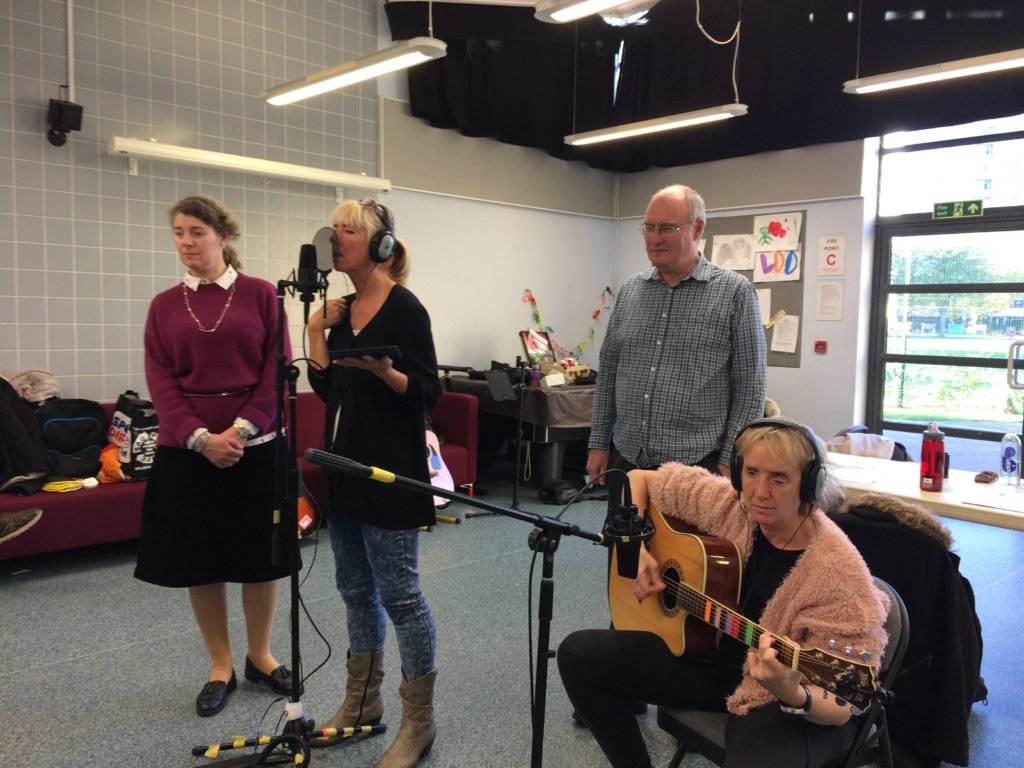 Included is a picture of some of the group recording their song. One participant is singing into the microphone, whilst another is sitting playing the acoustic guitar, which also has a microphone in front of it. Two other participants are standing listening.