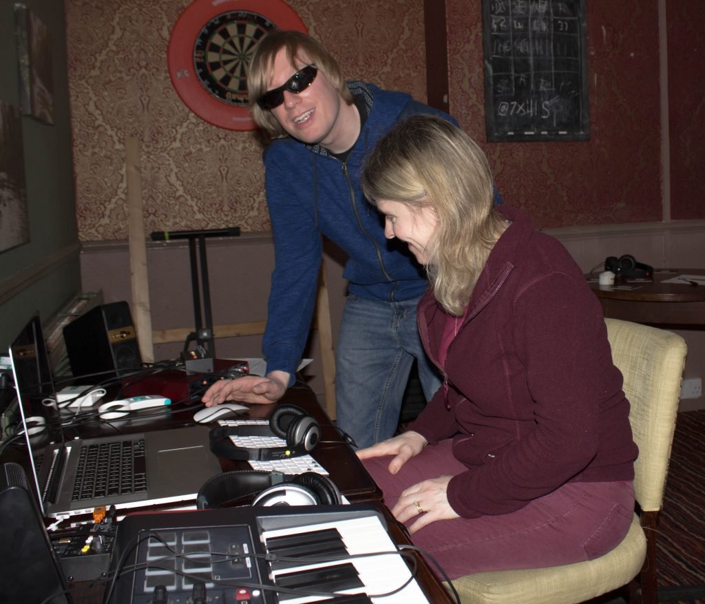 Included is a picture of participant Elena Sommers sitting with the laptop in front of her on a table. There is a MIDI Keyboard on her left and David standing to her right, with headphones on the table in front of them both. 