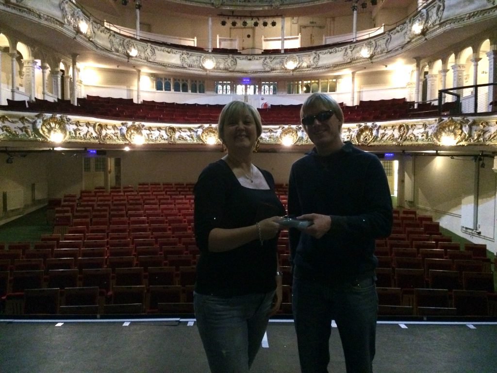 Rachel Goodall from The Red Sauce Theatre Company, and David Shervill from Global Music Visions C.I.C. standing on the stage at the New Theatre Royal, with their backs to the brightly lit auditorium. David is handing the CDs and memory stick to Rachel ready for their rehearsals.