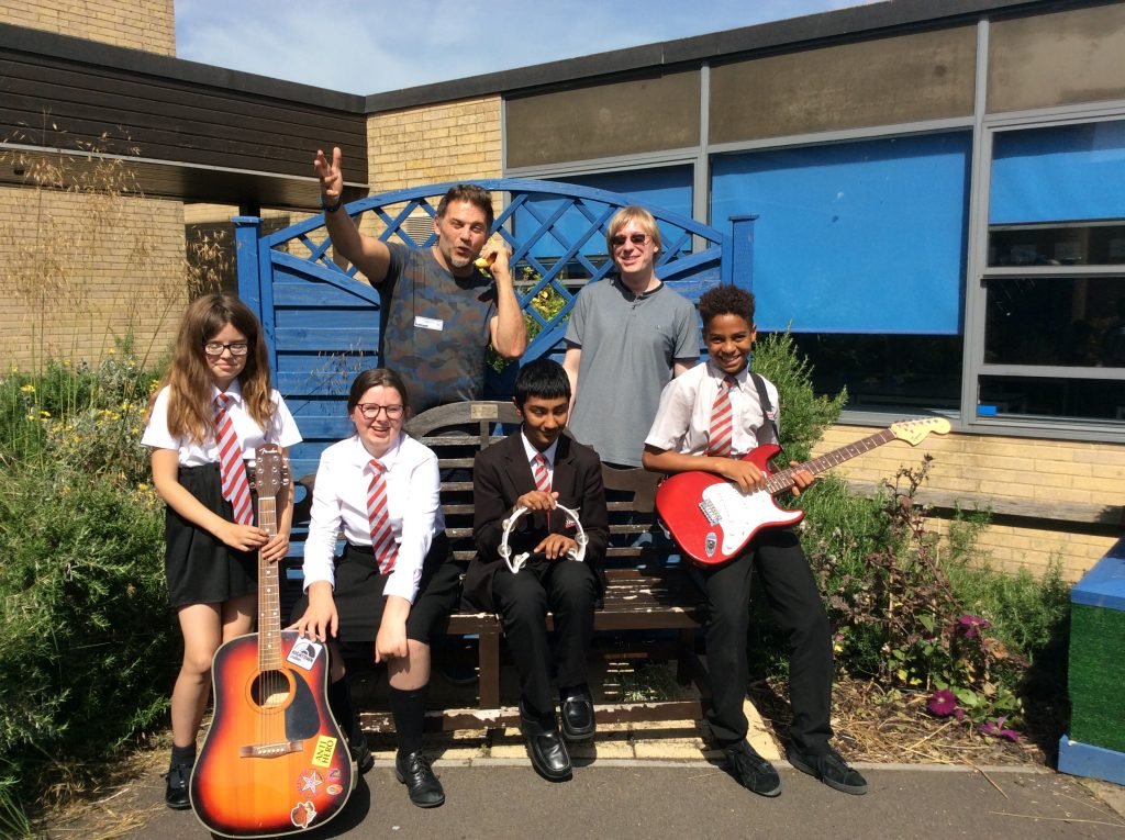 Included is a picture of some of the group sitting on the bench outside in the school gardens smiling. At either side of the bench one of the members is holding an acoustic guitar upright by their side, whilst another is holding an electric guitar across their lap. Two other students are sitting between them; one of them is holding a tambourine. David and Jim are standing behind the bench next to the green bushes.