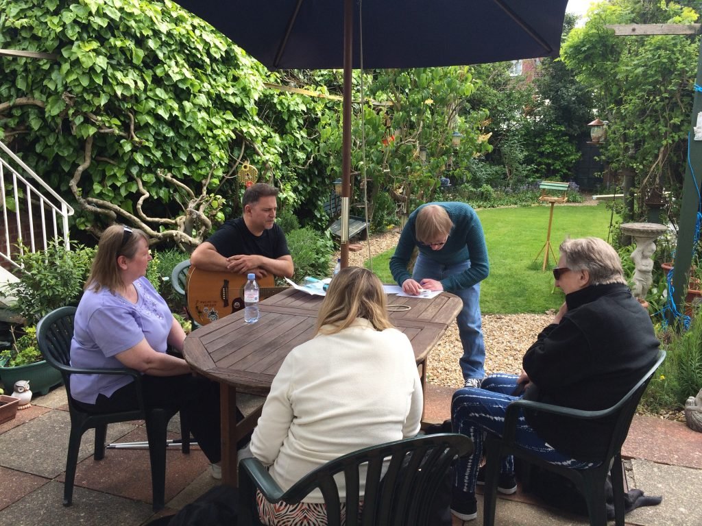 Included is a photo of five people sitting around a garden table outside, with trees and hedgerows around them. They are working through their lyrics, and one person is holding an acoustic guitar.