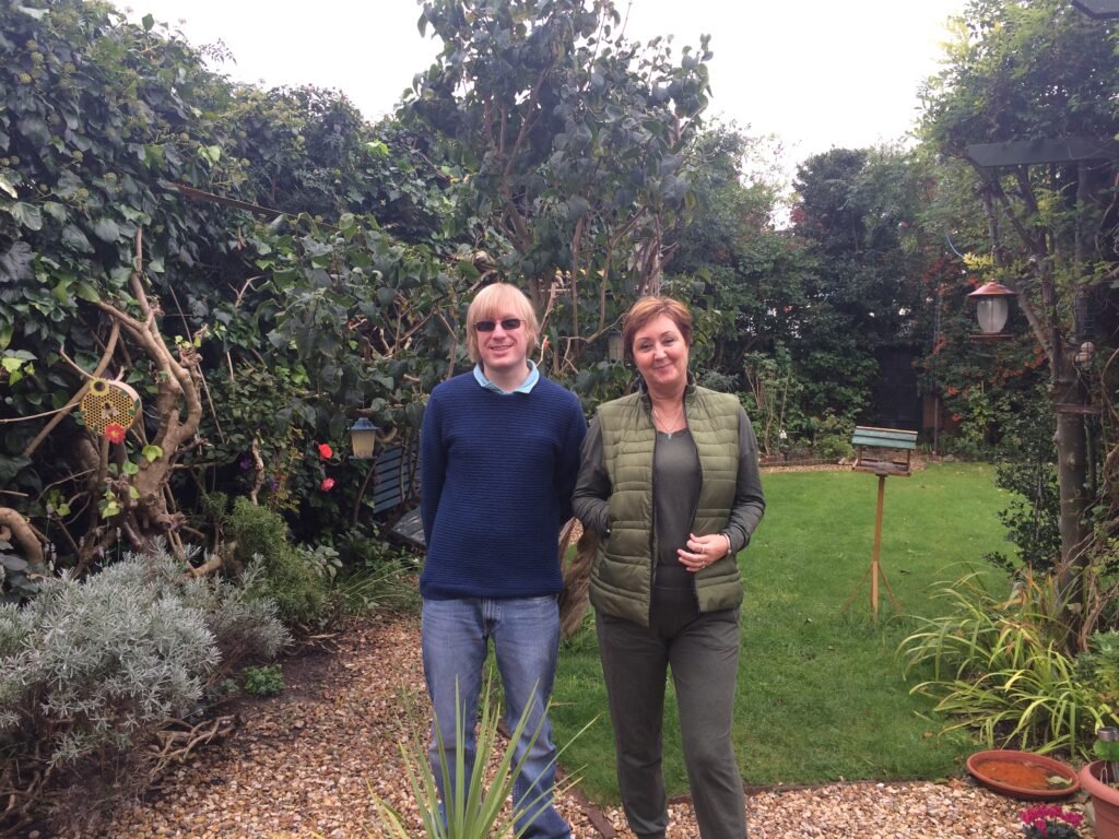 Photo of Rachel Goodall and David Shervill standing together on a shingle path, facing the camera. There are trees, hedgerows, and grass behind them.