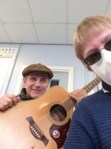 Included is a photo. As you look at the photo Jim Chorley is sitting to the left holding his acoustic guitar in front of him, and David Shervill who is wearing a face mask, is sitting to the right, both with some distance between them.