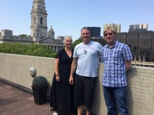 Included is a photo of 3 of the collaborative partners. They are standing side by side facing the camera, from left to right, Rachel Goodall from Red Sauce Inclusive Theatre Company, Billy Stevenson from Film Crew 4U CIC, and David Shervill from Global Music Visions C.I.C. The rooftop and Bell Tower of Portsmouth Guildhall can be seen in the background.