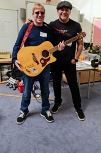 Image Description shows David Shervill and Jim Chorley facing the camera standing in front of the music equipment in the music room. David is holding an acoustic guitar, and there are percussion instruments behind them.