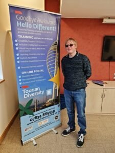 David is standing beside the Toucan Pull up Banner, and is facing the camera. Key information on the banner reads: Goodbye Awkward, Hello Different by Toucan Diversity. There is a picture of The Spinnaker Tower and Gunwharf Quays in the background. The Contact Number is 07834885539. 
