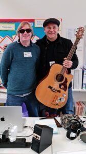 David Shervill and Jim Chorley are standing behind a table with a laptop on it, facing the camera. Jim is holding his acoustic guitar in his left hand.