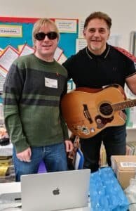 Photo shows David Shervill and Jim Chorley both smiling and facing the camera. They are standing behind a table with a laptop on it, and Jim is holding his acoustic guitar in front of him.