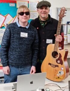 Photo shows David Shervill and Jim Chorley both smiling and facing the camera. They are standing behind a table with a laptop on it, and Jim is holding his acoustic guitar in his left hand.