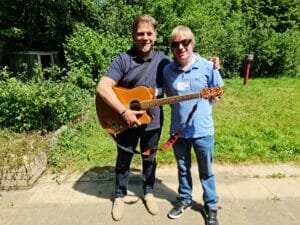 Jim and David are standing in the school grounds with grass and hedgerows behind them. They are facing the camera and Jim is holding his acoustic guitar.