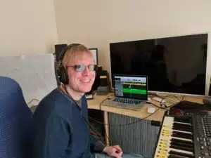 David Shervill (Founder) wearing headphones and sitting at a desk, with a laptop open directly in front of him. There are speakers, a sound card and a MIDI keyboard too.