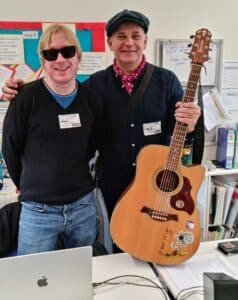 Photo shows David Shervill and Jim Chorley both smiling and facing the camera. They are standing behind a table with a laptop on it, and Jim is holding his acoustic guitar in his left hand.