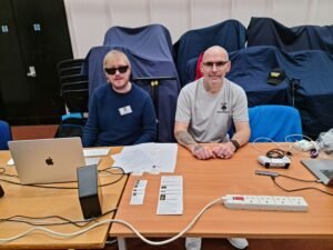 David and Sam are sitting at the table with their laptops, speakers and controllers positioned on the table. They are both facing the camera.